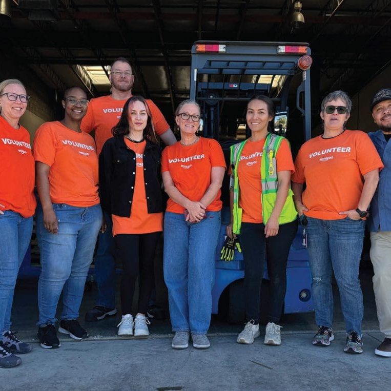 A group of eight volunteers in orange shirts stands together in front of a forklift in the pantry, ready to make a life-changing impact. Camille, at the forefront, beams with pride as they prepare for another day of service.