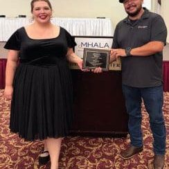 Two people stand indoors; the man on the right holds a Golden Bell Award plaque, and both are smiling at the camera. A sign behind them reads MHALA.