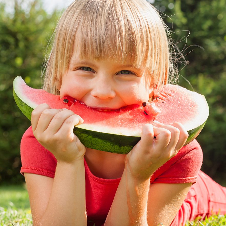 Happy little girl biting slice of melon lying in a summer garden