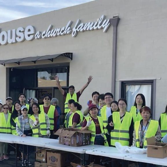 A group of people in yellow vests stand behind tables with boxes outside a building labeled The House, serving the community as a true church family.