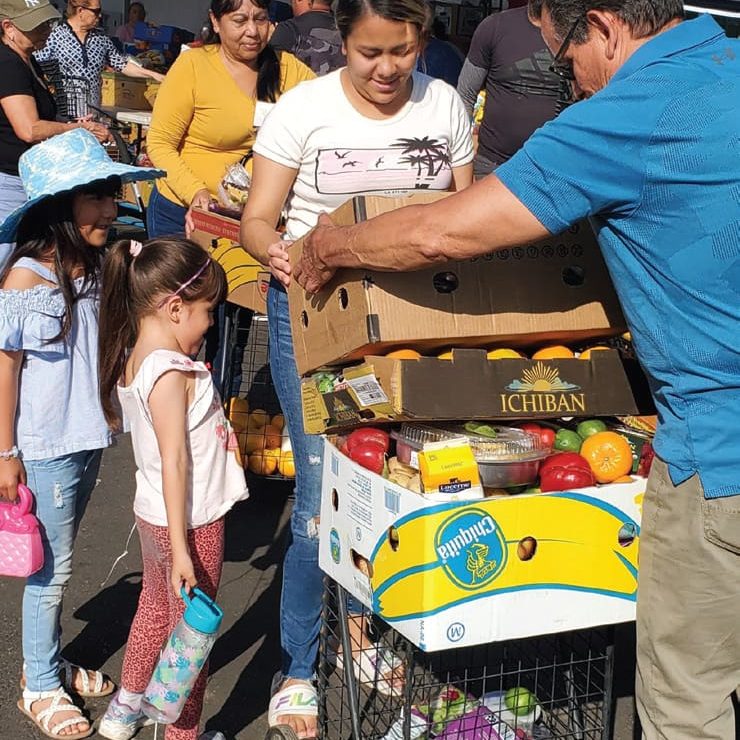 People gather around Camille's cart, filled with boxes of life-changing produce, at the bustling outdoor market.